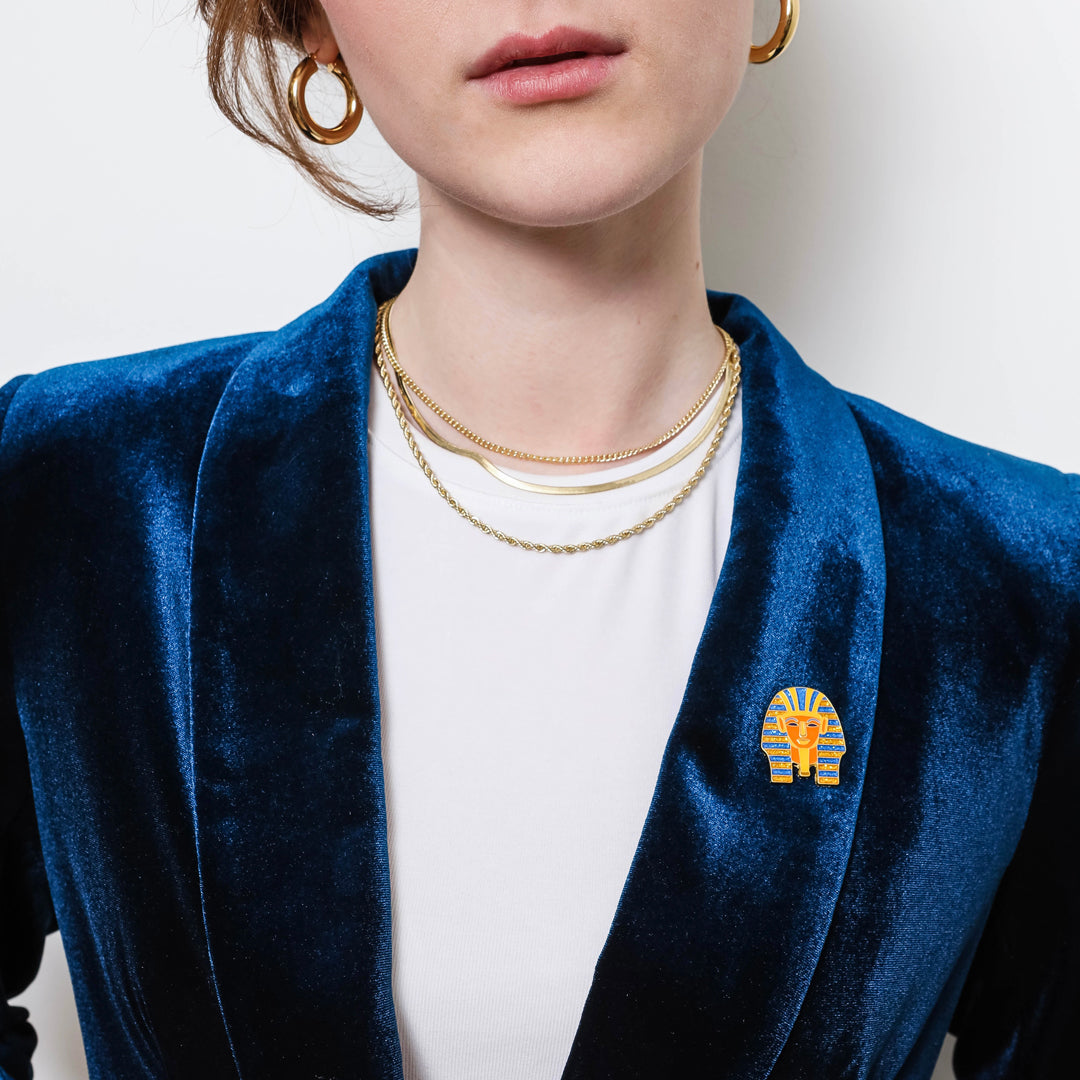 Person wearing a blue velvet blazer with a gold King Tut Lapel brooch, white shirt, and gold jewelry.