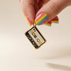 Hand holding a rainbow-colored cassette tape charm against a beige background