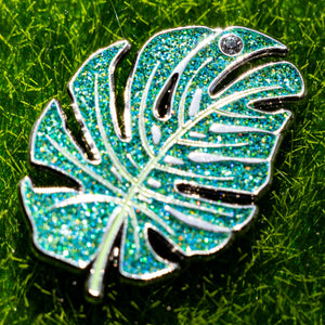 Decorative Monstera Leaf Enamel Pin with glittery turquoise and silver design on a grass background