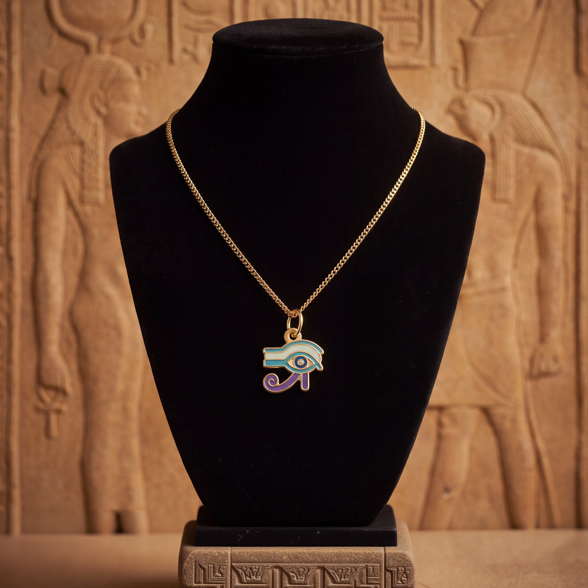 A gold chain necklace with an Egyptian Eye of Horus pendant, displayed on a black velvet jewelry bust against a warm, textured background resembling ancient stone carvings.