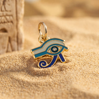 Gold-tone Egyptian Eye of Horus charm with blue and white enamel details, resting on sandy ground next to a blurred stone prop.