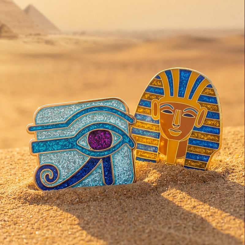 Two Egyptian-themed pins on sand with pyramids in the background