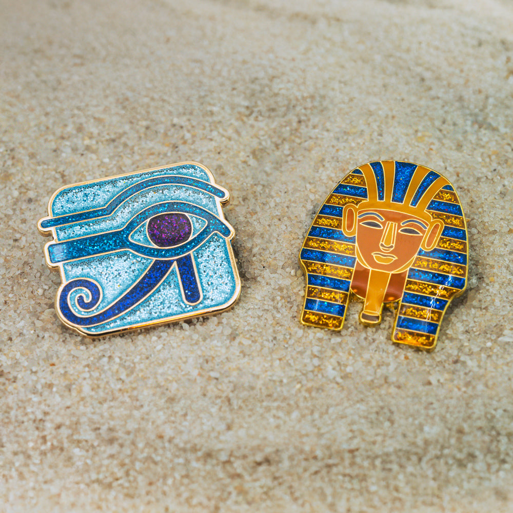 Two Egyptian-themed pins on a sandy background