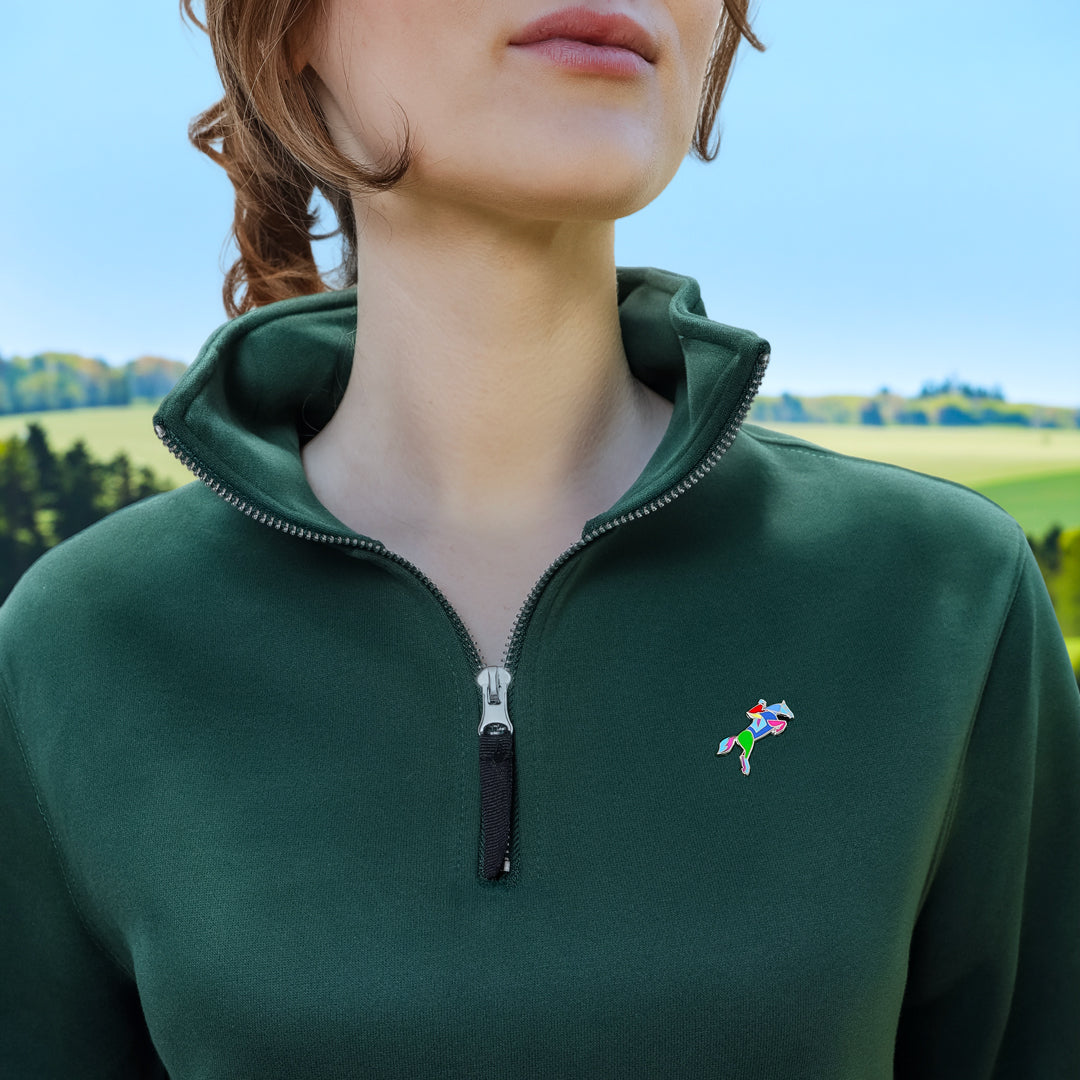 A close-up of a woman wearing a dark green quarter-zip pullover sweatshirt with a small colorful Horse Jumping Enamel Pin from Spot Studio Art on the left chest, set against a blurred outdoor landscape background.