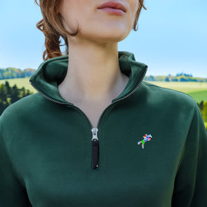 A close-up of a woman wearing a dark green quarter-zip pullover sweatshirt with a small colorful Horse Jumping Enamel Pin from Spot Studio Art on the left chest, set against a blurred outdoor landscape background.