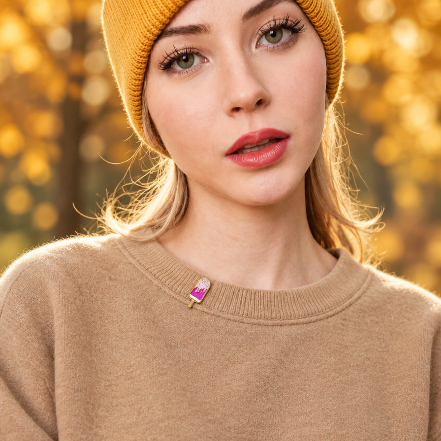A close-up of a woman wearing a tan knit crewneck sweater and a mustard yellow beanie, with a small colorful ice cream cone brooch pinned to her sweater at the collar. The background is a warm, blurred autumn park scene.