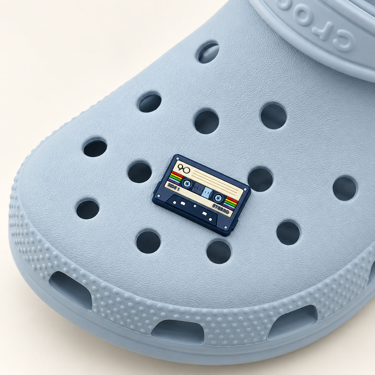 Navy cassette tape shoe charm with a cream label and rainbow accents attached to a powder-blue Croc.