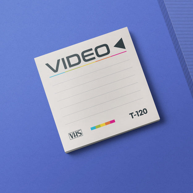 A square white VHS-style notepad or memo pad with the word VIDEO across the top, a small VHS logo, and the text T-120, placed on a blue background with a ribbed blue panel at right.