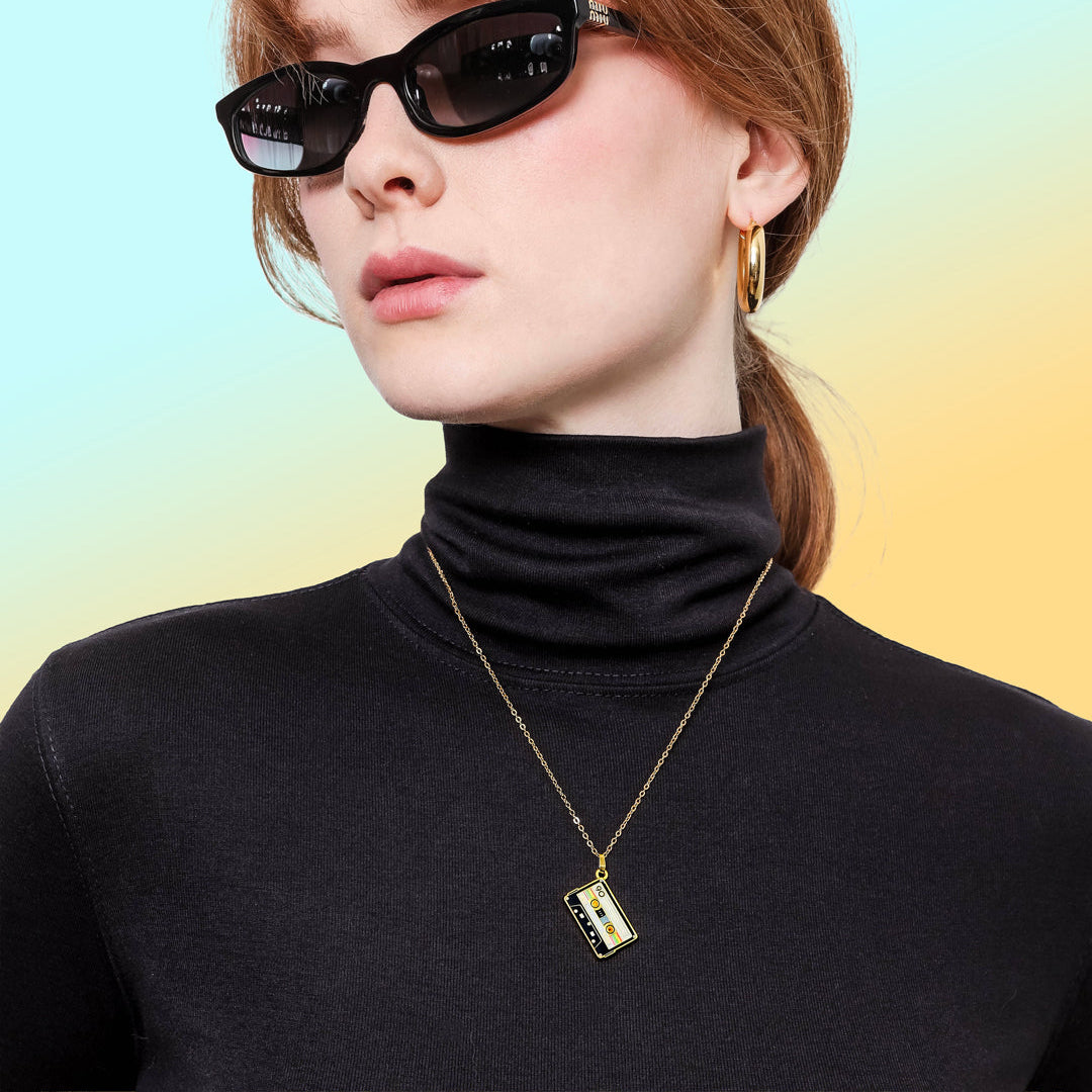 Woman wearing sunglasses and a black turtleneck with a gold Retro Cassette Tape Necklace from Spot Studio Art against a gradient background.
