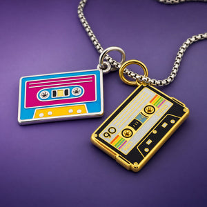 Two cassette tape pendant necklaces on a silver chain, one with a blue, pink, and yellow design and the other with a gold and black design, displayed on a purple background.