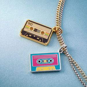 Two cassette tape pendant necklaces on gold and silver chains, displayed on a textured light blue background.