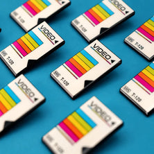 Several retro-style VHS Tape Enamel Pins arranged on a bright blue background. The cards are white with a colorful striped pattern in yellow, pink, and blue, and the word 'VIDEO' is printed on them.