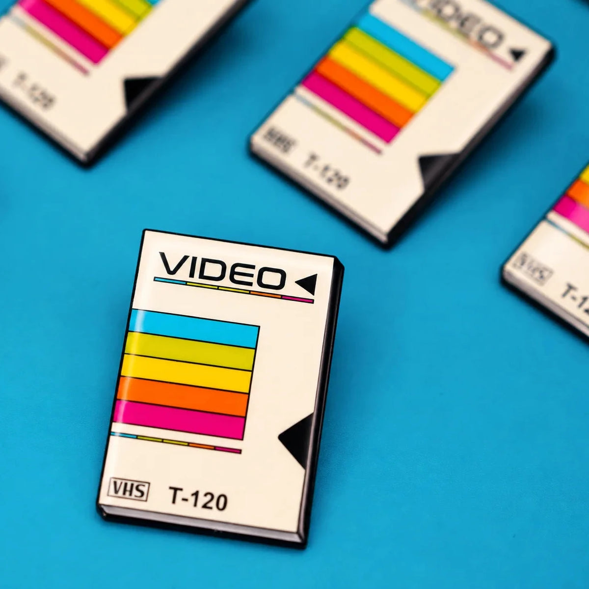 A close-up of a VHS-style cassette-shaped enamel pin with the word VIDEO and model text T-120, featuring a retro multicolor stripe graphic, placed on a bright blue background with several similar items blurred in the background.