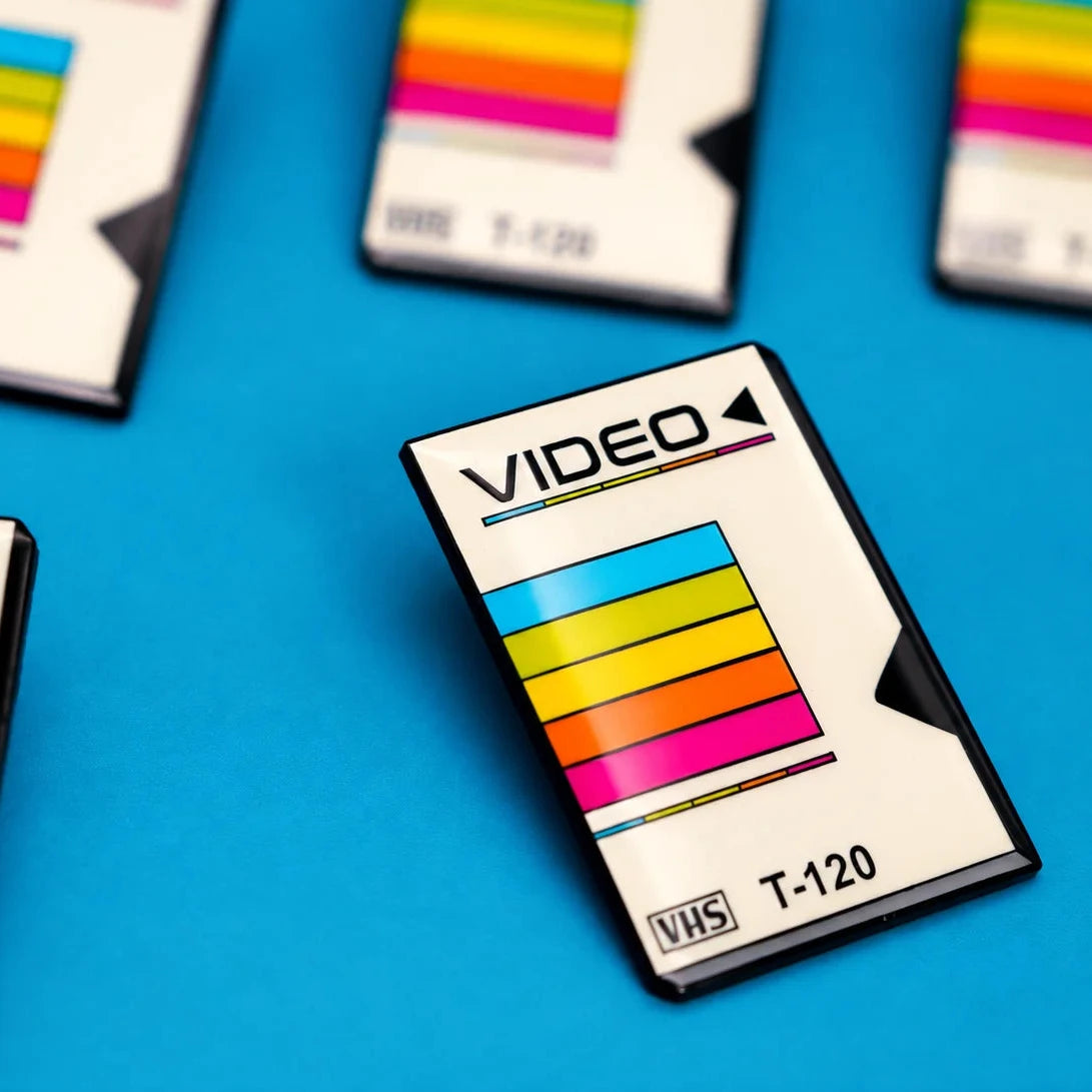 A close-up of a VHS tape enamel pin on a bright blue surface, with several similar cartridges partially visible in the background. The cartridge is white with a black plastic border and a colorful striped label featuring cyan, yellow, orange, and pink bands.