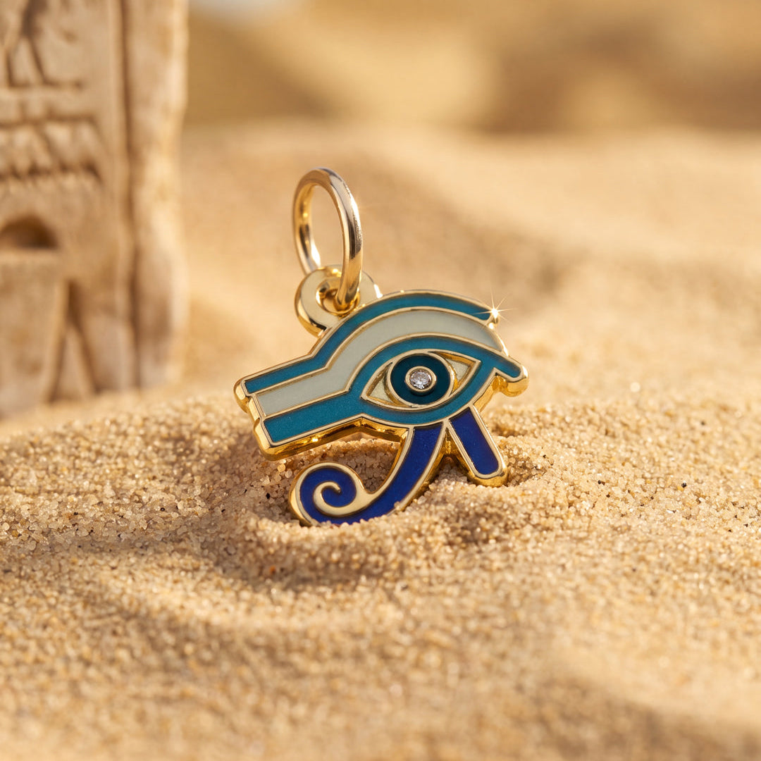 Gold-tone Egyptian Eye of Horus charm with blue and white enamel details, resting on sandy ground next to a blurred stone prop. 