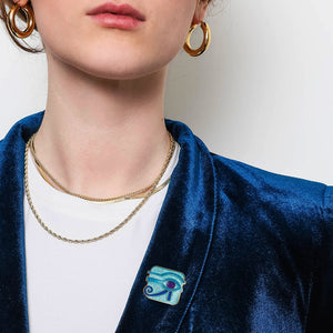 Person wearing a blue velvet blazer with a Eye of Horus lapel pin, white shirt, and gold jewelry on a plain background