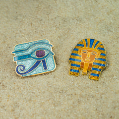 Two Egyptian-themed pins on a sand surface