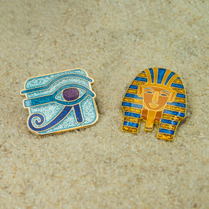 Two Egyptian-themed pins on a sand surface