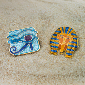 Two Egyptian-themed pins on a sandy background