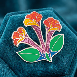 A colorful Honeysuckle enamel pin with green leaves, displayed on a textured teal velvet surface.