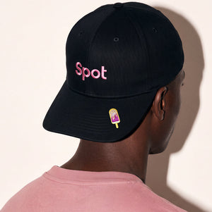 A man wearing a black baseball cap from the back, featuring the word 'Spot' embroidered in pink on the front and a small popsicle patch on the brim. He is wearing a dusty pink t-shirt against a warm off-white background.