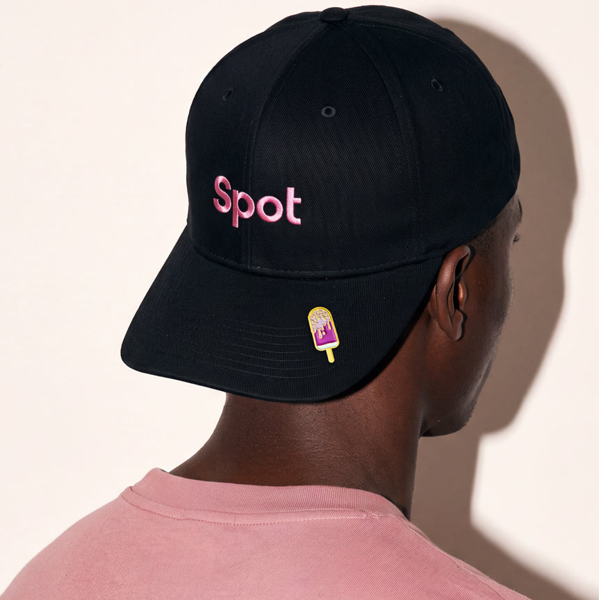 A man wearing a black baseball cap from the back, featuring the word 'Spot' embroidered in pink on the front and a small popsicle patch on the brim. He is wearing a dusty pink t-shirt against a warm off-white background.
