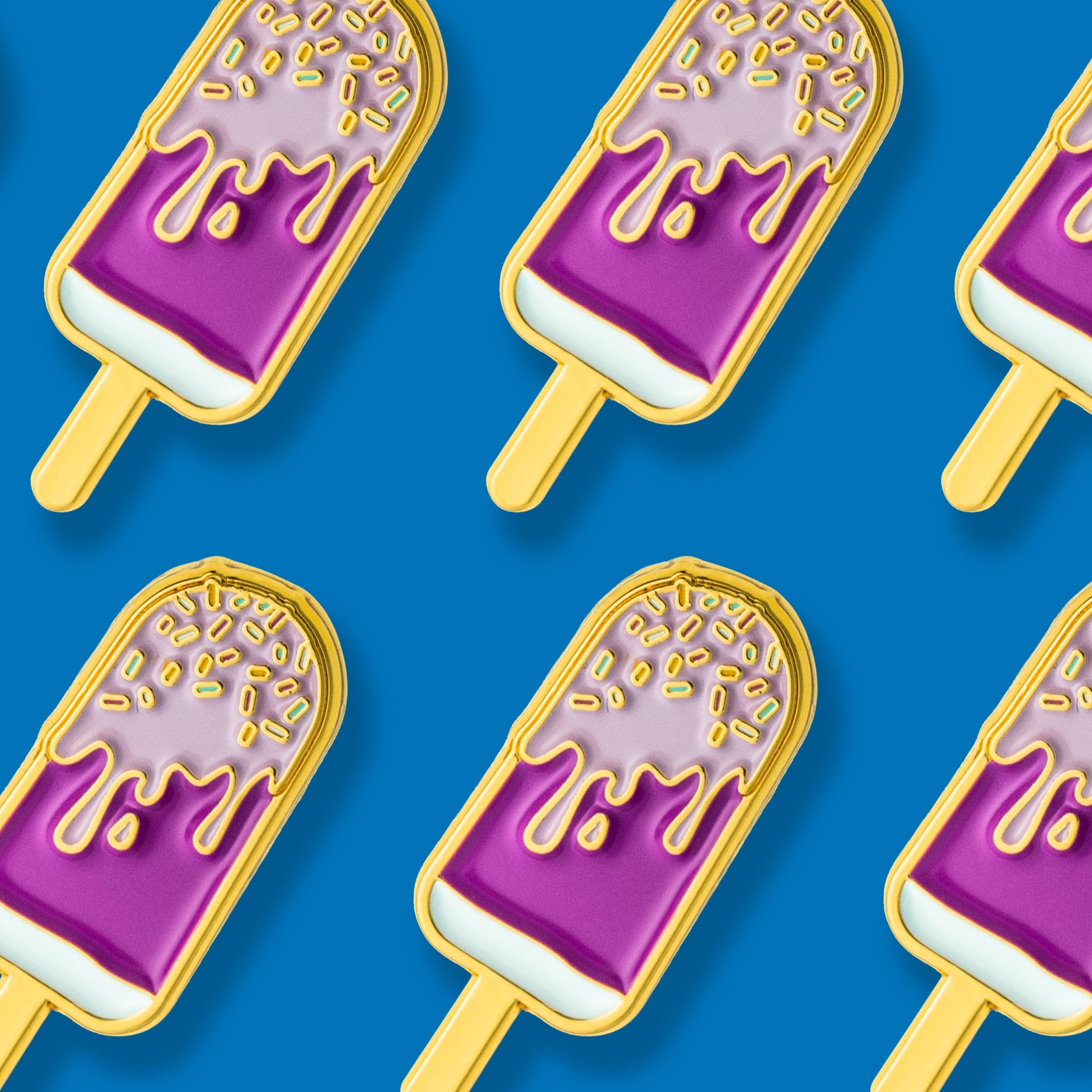 A series of gold-plated enamel pins shaped like melting ice cream popsicles with purple 'ice cream' and golden sprinkles on a blue background.