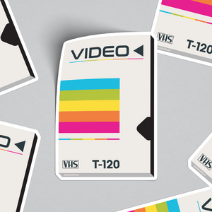 A collection of vinyl stickers designed to resemble vintage VHS tape sleeves, with a rainbow-colored stripe on each. The stickers are displayed on a neutral background.