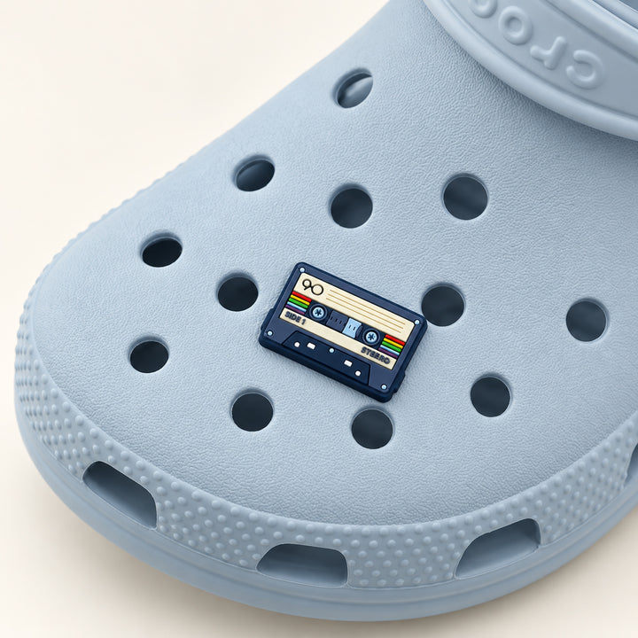Navy cassette tape shoe charm with a cream label and rainbow accents attached to a powder-blue Croc.
